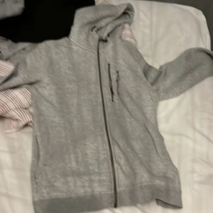 Grå huvtröja från Sail Racing - Säljer en grå huvtröja från Sail Racing med dragkedja och broderad logga. Tröjan har en bekväm passform och är perfekt för kyliga dagar. Den har en praktisk bröstficka med dragkedja och en mysig huva.