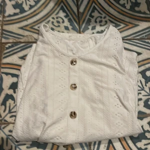 Vit blus med knappar - Säljer en stilren vit blus med dekorativa knappar framtill. Blusen har ett diskret hålmönster och långa ärmar, perfekt för en elegant look.