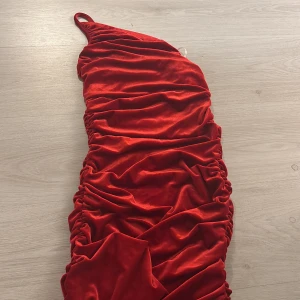 Röd oneshoulder klänning i sammet - Snygg röd oneshoulder klänning i mjuk sammet med rynkade detaljer längs sidorna. Perfekt för en kväll ute eller en speciell tillställning. Klänningen har en figurnära passform som framhäver silhuetten.