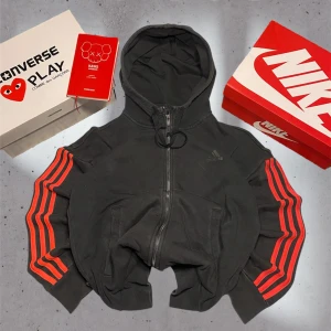 Svart hoodie från Adidas med röda ränder - Snygg svart hoodie från Adidas med ikoniska röda ränder längs ärmarna. Den har en dragkedja framtill och en praktisk huva. Perfekt för en sportig look.