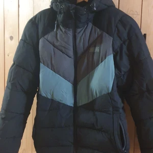 Svart och blå dunjacka från Urberg - Säljer en snygg och varm dunjacka från Urberg i svart med blå detaljer. Jackan har en huva och dragkedja framtill, perfekt för kyliga dagar. Den är designad med ett stilrent mönster och passar bra för vintern.