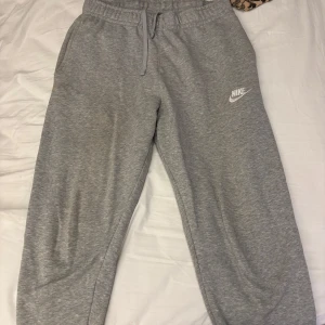 Grå mjukisbyxor från Nike - Säljer ett par grå mjukisbyxor från Nike med elastisk midja och snörning. Byxorna har en avslappnad passform och en bakficka. Perfekta för en chill dag hemma eller en snabb tur till affären.