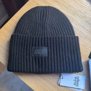Svart ribbad mössa från Twist & Tango - Snygg svart ribbad mössa från Twist & Tango. Perfekt för kyliga dagar med stilren design och ett diskret märke framtill. Passar både till vardags och mer uppklädda tillfällen.