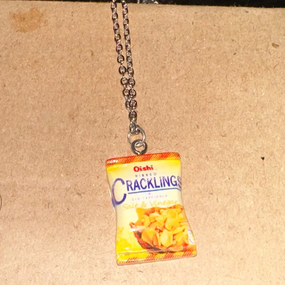 Unikt halsband med en rolig berlock som ser ut som en påse Oishi Cracklings. Perfekt för dig som vill ha något annorlunda och kul i din smyckessamling. Kedjan är silverfärgad och berlocken är färgglad med detaljer i gult och orange.. Accessoarer.