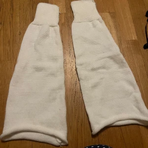 Vita stickade benvärmare - Säljer ett par vita stickade benvärmare. Perfekta för att hålla benen varma under kyliga dagar. De har en enkel design med ribbstickade kanter upptill och nedtill.