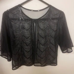 Svart transparent bolerotopp med paljetter - Elegant svart bolerotopp i transparent material med glittrande paljettdetaljer. Toppen har en öppen design framtill och trekvartsärmar. Väldigt fin att ha över tex ett linne storlek S/M 