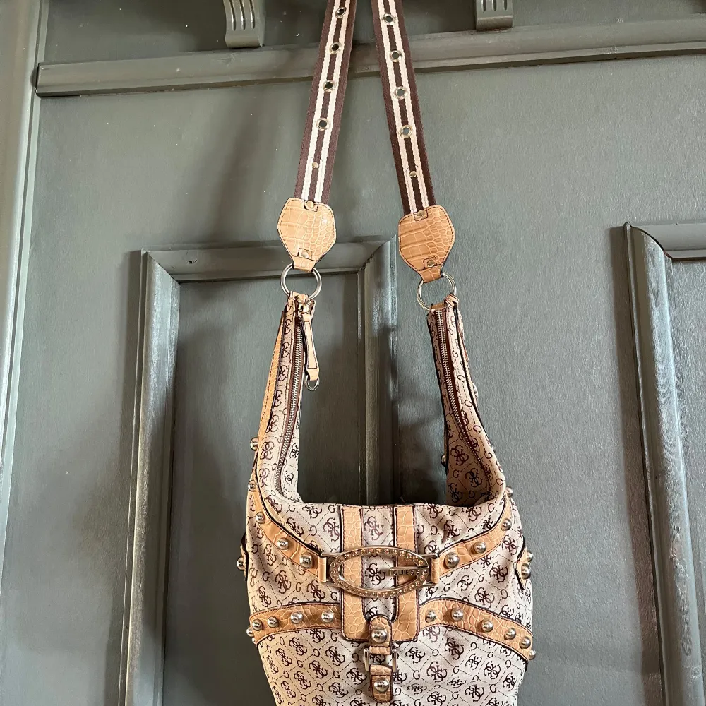 Snygg beige axelväska från Guess med monogrammönster och dekorativa nitar. Väskan har detaljer i syntetmaterial som ger en lyxig känsla. Perfekt för att ge din outfit en extra touch. Väskan är endast använd ett fåtal gånger, därefter endast förvarats i garderoben. Finns ett par fläckar i innefoderet. . Laukut & Käsilaukut.