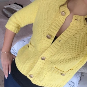 Gul stickad kofta med knappar - Säljer en solgul stickad kofta med trekvartsärmar och stora knappar framtill. Koftan har två fickor och ribbade detaljer vid ärmslut och nederkant. Perfekt för att lysa upp din garderob!