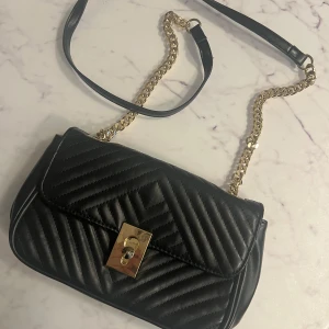 Svart quiltad axelväska med guldkedja - Snygg svart axelväska i quiltat mönster med guldkedja och låsdetalj. Perfekt för att ge en elegant touch till din outfit. Väskan har en praktisk storlek och en stilren design.