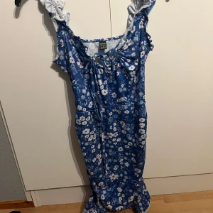 Blå blommig klänning från Dreimp - Charmig blå klänning med blommigt mönster från Dreimp. Klänningen har volangdetaljer vid axlarna och en knytning framtill. Perfekt för en somrig look!