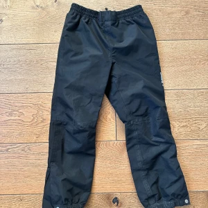 Svarta byxor med resårmidja - Säljer ett par svarta byxor med resårmidja och justerbara benslut. Perfekta för utomhusaktiviteter med slitstarkt material och förstärkta knän. Praktiska och bekväma för alla väder.