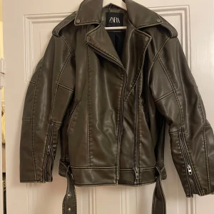 Mörkgrön skinnjacka från Zara - Snygg grön skinnjacka från Zara med dragkedja och knappar. Jackan har en klassisk bikerstil med bälte i midjan och långa ärmar. Perfekt för en cool look.