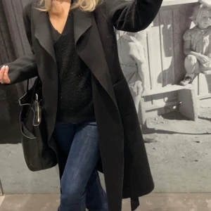 Svart kappa från Vila - Elegant svart kappa från Vila med en stilren design. Perfekt för att hålla sig varm och snygg under kyligare dagar. Kappan har en klassisk passform och är ett mångsidigt tillskott till garderoben.