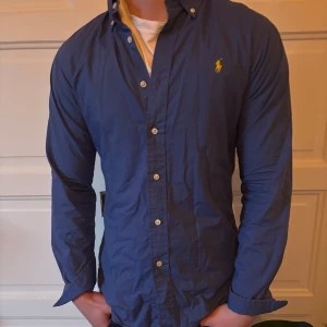 Blå skjorta från Polo Ralph Lauren - Snygg blå skjorta från Polo Ralph Lauren i slim fit. Skjortan har långa ärmar och knappar framtill. Den är dekorerad med det klassiska broderade logotypen i gult på bröstet.