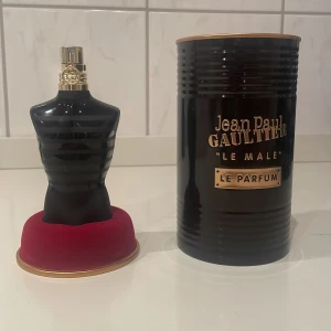 Jean Paul Gaultier Le Male Le Parfum - Säljer en elegant parfymflaska formad som en manlig torso i svart glas med gulddetaljer. Flaskan står på en röd bas och kommer med en stilren svart metallförpackning. Doften är intensiv och maskulin, perfekt för den som vill sticka ut. Det finns ungefär kvar 70ml i flaskan utav 75ml.