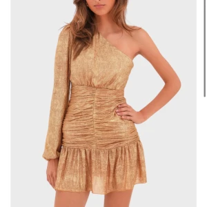 Guldig oneshoulder klänning - Trendig guldglittrig klänning med oneshoulder-design och lång ärm. Den har en åtsittande passform med rynkade detaljer i midjan och en volangkant nedtill. Perfekt till nyår 🤗