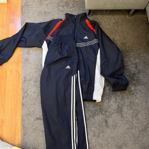 Marinblått träningsset från Adidas - Säljer ett marinblått träningsset från Adidas med vita och röda detaljer. Setet består av en jacka med dragkedja och matchande byxor. Perfekt för träning eller en avslappnad dag. Vindklädes material. Aldrig använda 