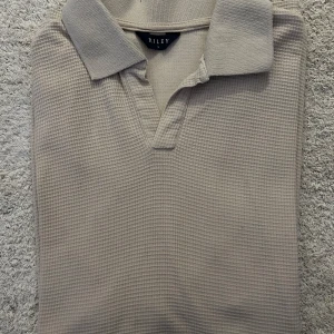 Beige pikétröja från Riley - Snygg beige pikétröja från Riley med klassisk krage och diskret textur. Perfekt för en stilren och avslappnad look.