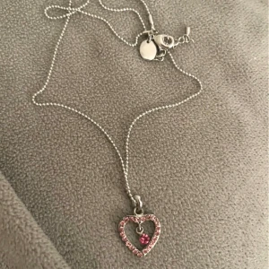 Halsband med hjärthänge och rosa sten - Nytt halsband med ett hjärta dekorerat med rosa stenar och en rosa pärla i mitten. Kedjan är i silverfärg och har ett justerbart lås.🩷