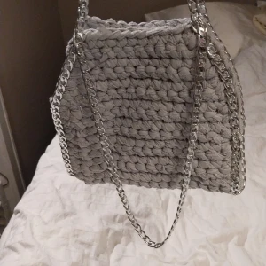 Grå virkad väska med kedjedetaljer - Snygg grå virkad väska med silverfärgade kedjor som axelrem. Perfekt för att ge din outfit en unik touch. Väskan har en mjuk och mysig känsla tack vare det stickade materialet.