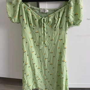 Grön blommig klänning från Chiquelle - Söt grön klänning med blommigt mönster från Chiquelle. Klänningen har korta puffärmar och en knytning framtill. Perfekt för somriga dagar med sin lätta och luftiga design.