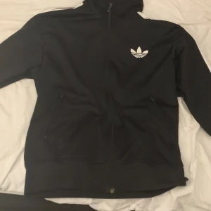 Svart jacka från Adidas - Säljer en svart jacka från Adidas med dragkedja och det klassiska logotypen på bröstet. Jackan har långa ärmar och vita ränder längs axlarna. Perfekt för en sportig look.
