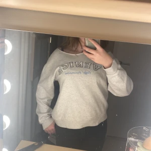 Grå sweatshirt med tryck - Säljer en grå sweatshirt med texten 'Venice' i blått och vitt tryck. Tröjan har en avslappnad passform och långa ärmar. Perfekt för en casual look.