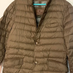 Brun dunjacka från Massimo Dutti - Säljer en stilren brun dunjacka från Massimo Dutti. Jackan har en quiltad design med knappar framtill och praktiska fickor. Perfekt för kyligare dagar.