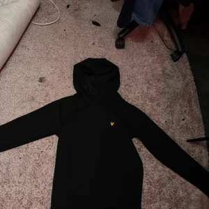 Svart hoodie från Lyle & Scott - Säljer en stilren svart hoodie från Lyle & Scott med en liten gul logga på bröstet. Perfekt för en avslappnad look. Hoodien har långa ärmar och en klassisk huva. Ord 700kr