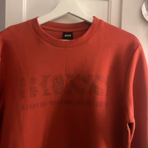 Röd sweatshirt från Boss - Snygg röd sweatshirt från Boss med ett subtilt mönster av märkesnamnet på bröstet. Tröjan har långa ärmar och en rund halsringning. Perfekt för en avslappnad stil.