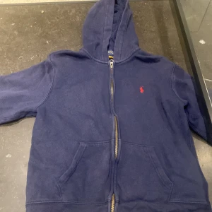 Mörkblå hoodie från Ralph Lauren - Säljer en snygg mörkblå hoodie från Ralph Lauren med dragkedja och huva. Den har en liten röd logotyp på bröstet och praktiska fickor framtill. Perfekt för en avslappnad stil.