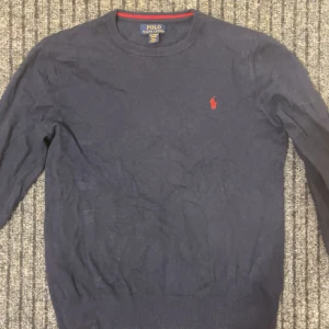 Mörkblå tröja från Polo Ralph Lauren - Säljer en stilren mörkblå tröja från Polo Ralph Lauren i Pima Cotton. Tröjan har en klassisk design med långärmade ärmar och en liten broderad logotyp i rött på bröstet. Perfekt för en avslappnad men ändå elegant look.