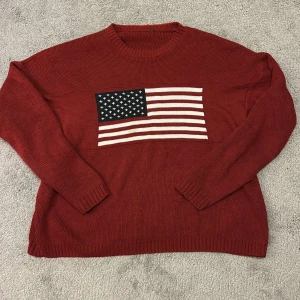 Röd stickad tröja med amerikansk flagga - Säljer en röd stickad tröja med en stor amerikansk flagga på framsidan. Tröjan har långa ärmar och en rund halsringning. Perfekt för en avslappnad stil.