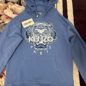 Blå hoodie från Kenzo - Snygg blå hoodie från Kenzo med det ikoniska tigertrycket och texten 'PARIS' på framsidan. Tröjan har en justerbar huva med snören och en praktisk magficka. Perfekt för en avslappnad stil.