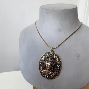  guldhalsband med dekorativt hänge - Unikt halsband i antik stil med ett stort, detaljerat hänge föreställande ett ansikte omgivet av blommönster. Kedjan är tunn och elegant i guldton. Perfekt för att ge en bohemisk touch till din outfit.