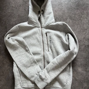 Grå hoodie från Sail Racing - Säljer en grå hoodie från Sail Racing med dragkedja och justerbar huva. Tröjan är i toppskick i storlek s och orginalpriset ligger på 1000kr men säljer denna för 350 då den aldrig används, tveka inte att höras av vid intresse😊