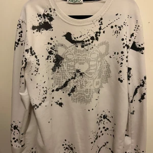 Vit tröja med tigertryck från Kenzo - Säljer en vit tröja från Kenzo med ett coolt tigertryck i svart och grått. Tröjan har ett unikt stänkmönster och långa ärmar. Perfekt för dig som vill sticka ut med stil!