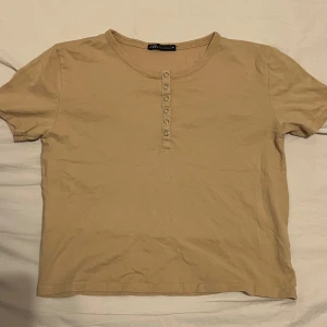 Beige kortärmad topp från Zara - Säljer en beige kortärmad topp från Zara med knappar framtill. Perfekt för en avslappnad stil. Toppen är i ett mjukt material och har en rund halsringning.