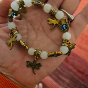 Armband med vita pärlor och berlocker - Charmigt armband med vita pärlor och antikfärgade berlocker i form av trollsländor och hamsa-händer med blå detaljer. Perfekt för att ge en bohemisk touch till din stil.