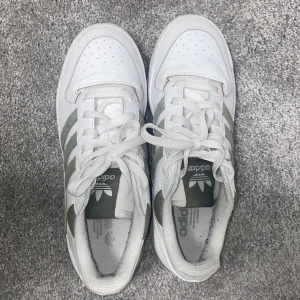 Vita Adidas sneakers med grå detaljer - Snygga vita Adidas sneakers med klassiska tre ränder i grått på sidan. Skorna har snörning och perforerade detaljer på tån för extra stil. Perfekta för en sportig look. Nästan helt nya bara använd 1 gång. Priset kan diskuteras