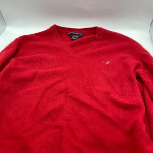 Röd ulltröja från GANT - Säljer en snygg röd tröja från GANT i 100% Geelong lammull. Tröjan har en v-ringad design och är perfekt för kyligare dagar. Den är både stilren och bekväm, vilket gör den till ett utmärkt val för höst och vinter. Den har krympt i tvätten så sitter som M även fast det står xl, om man är intresserad av hur den sitter så skickar jag gärna en bild.