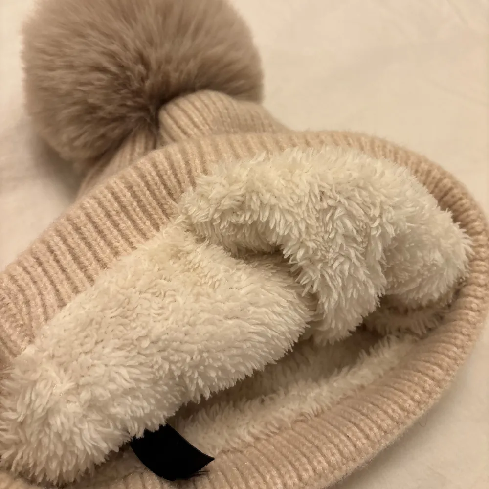 Mysig beige stickad mössa med en fluffig pälsboll på toppen. Insidan är fodrad med mjuk fleece för extra värme. Perfekt för kalla vinterdagar.Mössan har aldrig använts eftersom jag inte passar i mössor.. Asusteet.