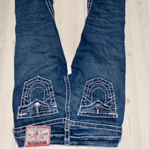 Blå jeans från True Religion - Säljer ett par blå jeans från True Religion med vita detaljer och kontrastsömmar. Byxorna har låg midja och är bootcut