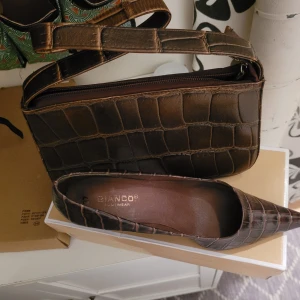 Bruna pumps från Bianco - Eleganta bruna pumps från Bianco med krokodilpräglat mönster. Skorna har en spetsig tå och hög klack, vilket ger en stilren look. Perfekta för att lyfta din outfit, med liknande väsk