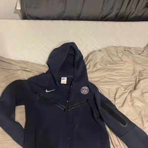 Mörkblå hoodie från Nike x PSG - Säljer en snygg mörkblå hoodie från Nike i samarbete med Paris Saint-Germain. Den har en dragkedja framtill och PSG-loggan på bröstet. Perfekt för fotbollsfans! Hoodien är i bra skick och har långa ärmar med en cool ficka på ärmen. Passar perfekt för höst och vår.