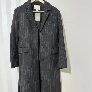 Grå randig kappa från H&M - Säljer en stilren grå kappa från h&m i storlek S . Den har ett klassiskt randigt mönster och är utrustad med knappar framtill och ett bälte i midjan för en snygg passform. Perfekt för höst och vinter, med långa ärmar och en elegant look.