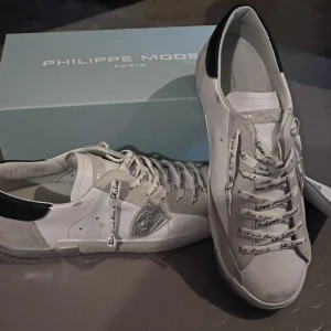 Vita sneakers från Philippe Model - Säljer ett par snygga vita sneakers från Philippe Model i skinn. De har coola detaljer som snörning med text och en svart häl. Perfekta för både vardag och fest! Kommer i originalkartong.