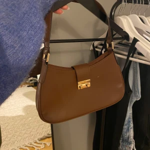 Brun axelväska med gulddetaljer - Säljer en snygg brun axelväska i skinn med gulddetaljer. Väskan har en stilren design med ett spänne framtill. Perfekt för både vardag och fest! Använd fåtal gånger!