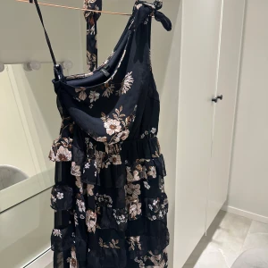 Blommig klänning från Vero Moda - Säljer en superfin blommig klänning från Vero Moda. Den är svart med vackra blommönster i ljusa toner. Klänningen har volanger och en oneshoulder-design som ger en unik look. Perfekt för sommarens fester eller en mysig vårdag. Materialet känns lätt och bekvämt. Passa på att fynda denna skönhet!