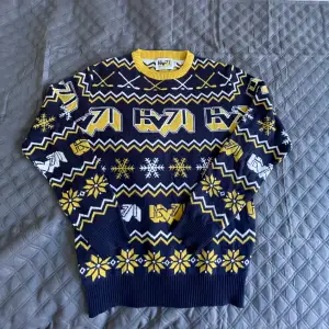 Säljer en supercool tröja från HV71 med ett snyggt mönster i blått, gult och vitt. Perfekt för vintern med långa ärmar och ett mysigt stickat material. Den har ett sportigt tema med hockeyklubbor och snöflingor. Grym för att visa ditt stöd för laget!